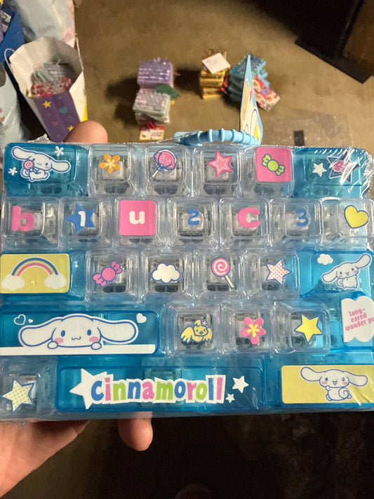 A hand holding a Cinnamoroll-themed plastic organizer tray with multiple small clear compartments decorated with colorful stickers and letter/number tiles, photographed indoors on a carpeted floor with other items in the background.
