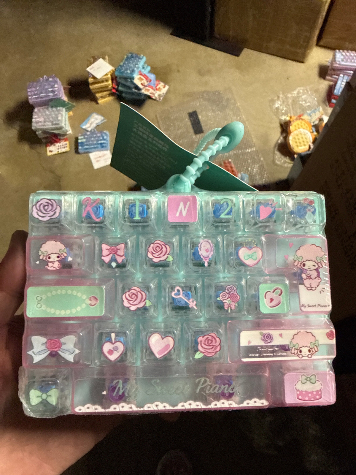 A hand holding a clear plastic organizer case filled with pastel decorative charms and stickers, including bows, hearts, flowers, and cartoon characters. The case has a mint green and pink color scheme with the text 'My Sweet Planner' visible on the front.