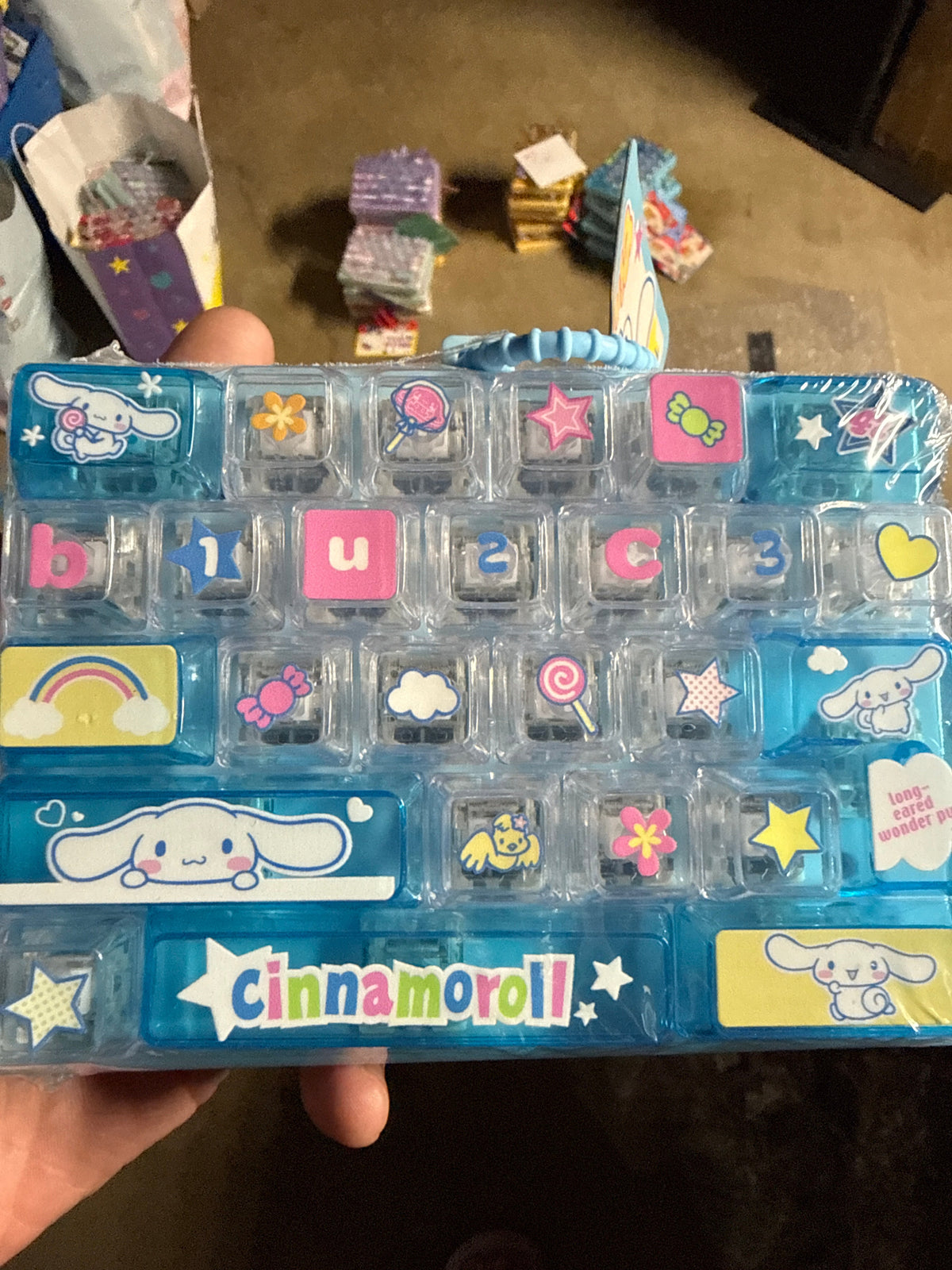 A hand holding a Cinnamoroll-themed plastic organizer tray with multiple small clear compartments decorated with colorful stickers and letter/number tiles, photographed indoors on a carpeted floor with other items in the background.